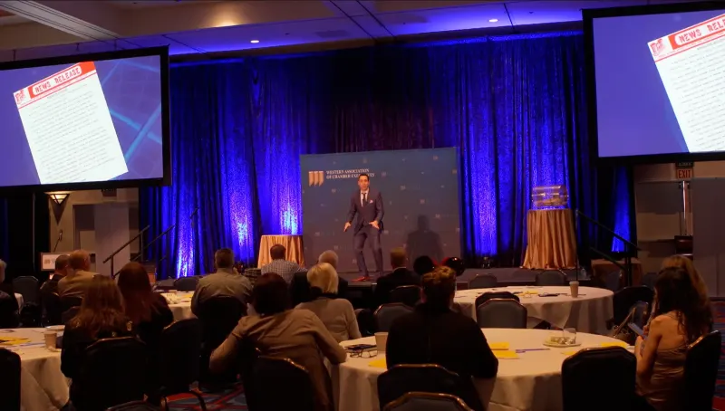 Jess Todtfeld on a ballroom stage under blue stage light, speaking to a conference audience seated at round tables