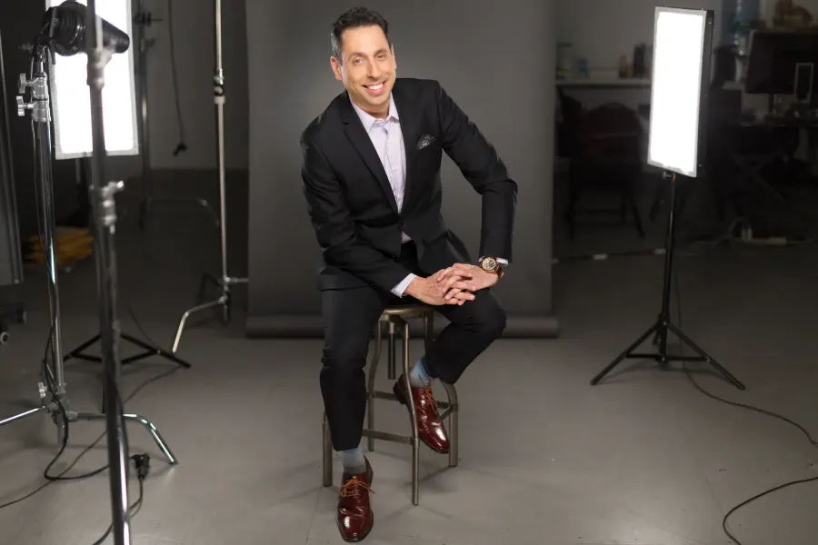 Jess Todtfeld seated on a stool between two studio lights, smiling, in a dark suit